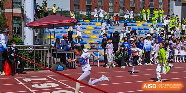 A young girl participating in an outdoor track and field event, running in a sack race in front of seated spectators in the stands, at Beijing SMIC Private School.