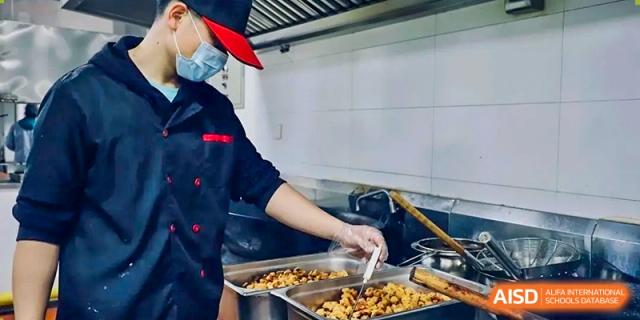 A cafeteria staff member at Boya International Academy (BIA) wearing a mask and uniform, serving food from a large warming tray in the kitchen.