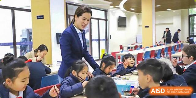 A female teacher from Boya International Academy (BIA) in a suit interacting with students seated at a dining table during a meal.