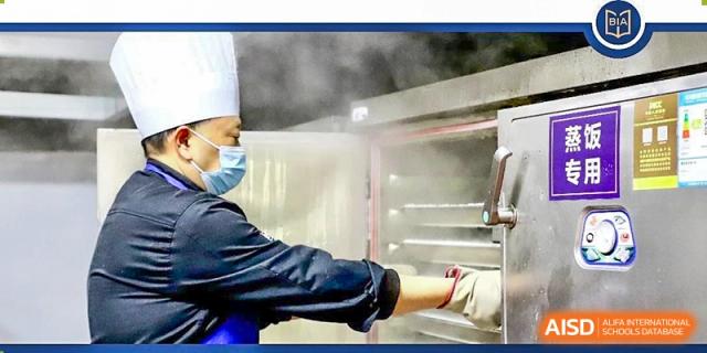 A chef from Boya International Academy (BIA) wearing a uniform and mask, removing food from a steaming oven in the school kitchen.