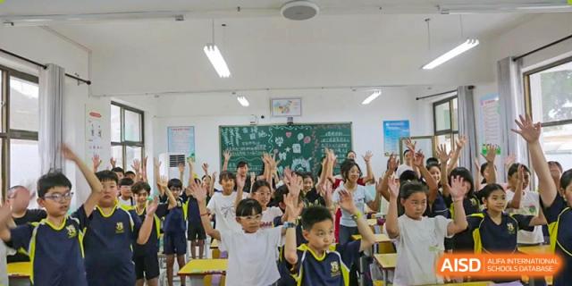 A large group of young students in a classroom raising their hands enthusiastically, showing high engagement, Brilliant Light School SJZ.