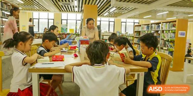 A teacher helping a group of students who are reading and studying together at a large table in the library, Brilliant Light School SJZ.