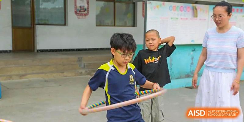 A male student in a blue and yellow uniform holding a wooden stick, with another student behind him, on a playground, Brilliant Light School SJZ.
