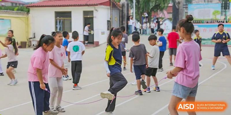 A female student in a yellow and blue uniform doing a high leg kick while jump roping with a group of other students in the background, Brilliant Light School SJZ.