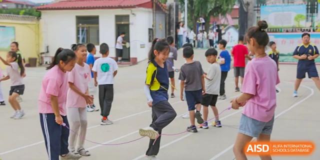 A female student in a yellow and blue uniform doing a high leg kick while jump roping with a group of other students in the background, Brilliant Light School SJZ.