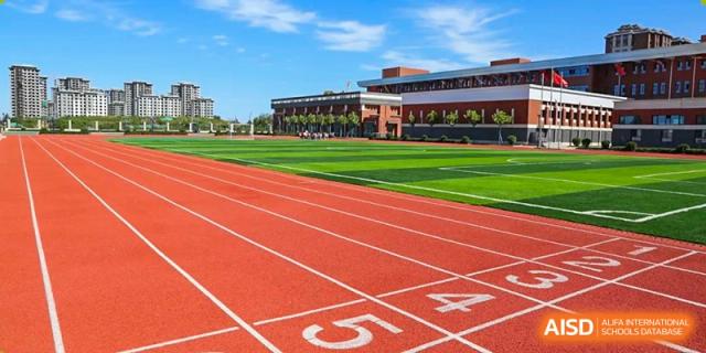 An outdoor running track with lane markings and a green sports field next to the modern school building under a bright blue sky, Brilliant Light School SJZ.