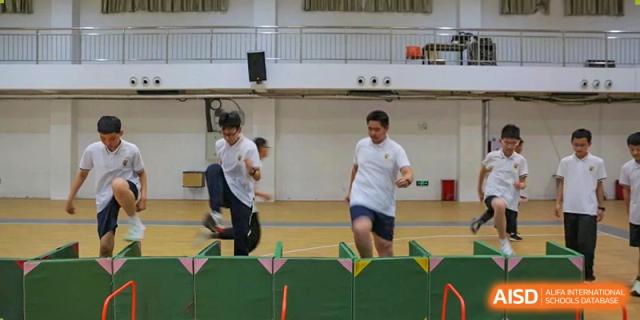 Two students in white t-shirts running and jumping over low green hurdles during a physical education class or sports practice, Brilliant Light School SJZ.