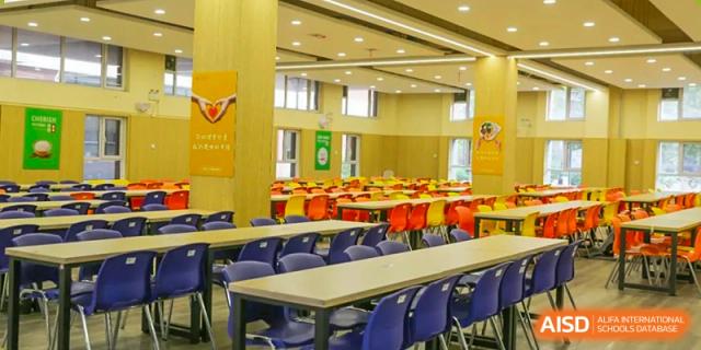 A large, brightly lit school cafeteria with rows of long tables and blue and purple chairs, Brilliant Light School SJZ.