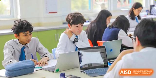 A male student in uniform sitting at a desk and working on a laptop computer in a modern classroom setting at Britannica International School, Shanghai.