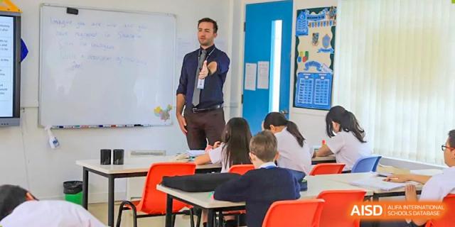A male teacher in a shirt and tie presenting and gesturing to a class of students seated at their desks in a classroom at Britannica International School, Shanghai.