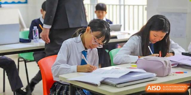 A female student in uniform focused on taking notes and studying a textbook at her desk in a classroom at Britannica International School, Shanghai.