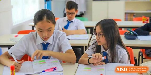Two female students in uniform collaborating on written work and assignments at their shared desk in a classroom at Britannica International School, Shanghai.