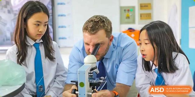 A male science teacher in a shirt and tie looking intently through a microscope in a laboratory or classroom at Britannica International School, Shanghai.