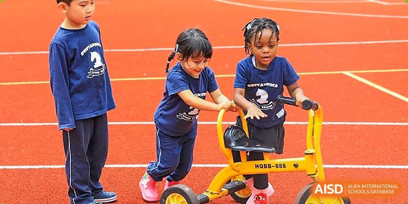 Two young children, a girl and a boy, playing on a yellow tricycle on a bright orange sports court at Britannica International School, Shanghai.