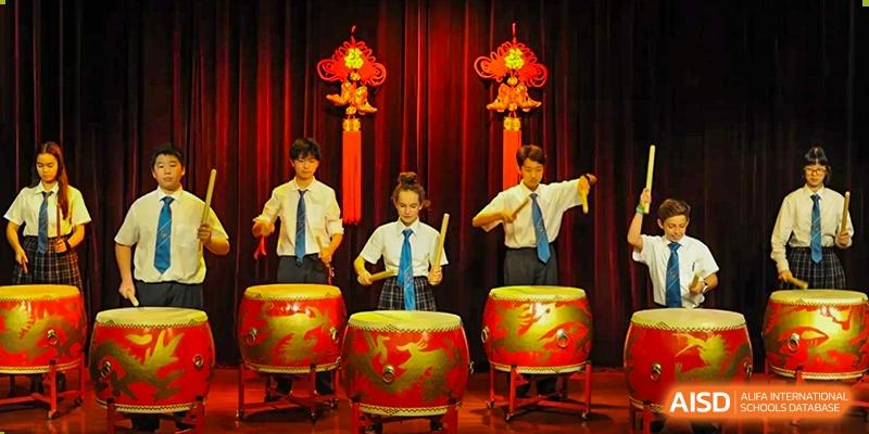 Four students in school uniform performing on large traditional Chinese drums under red decorations and lighting during a cultural event at Britannica International School, Shanghai.