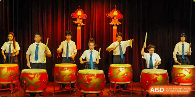Four students in school uniform performing on large traditional Chinese drums under red decorations and lighting during a cultural event at Britannica International School, Shanghai.