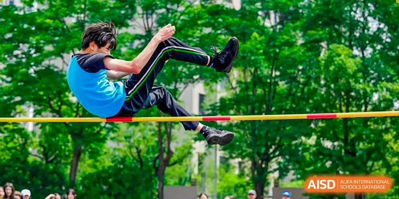 A close-up of a student in green uniform mid-air, attempting a high jump over the bar at Britannica International School, Shanghai.