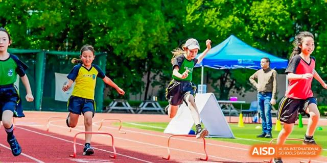 A student in a green cap and uniform leaping over an obstacle on an athletic track during a sports day event at Britannica International School, Shanghai.