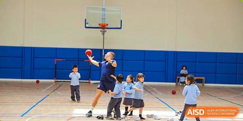 An older student shooting a basketball while younger students watch during a training session on the outdoor court at Britannica International School, Shanghai.