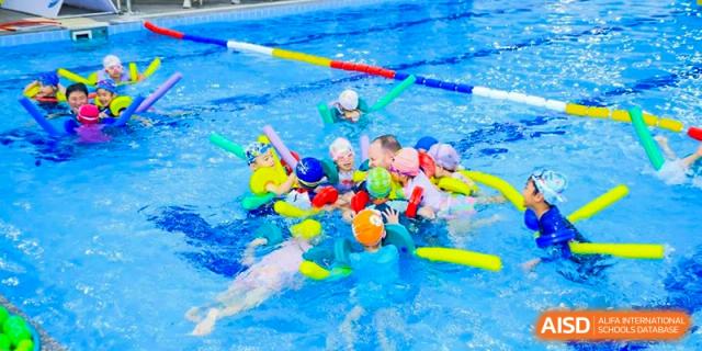 A group of young children wearing swim caps participating in a fun swimming activity or lesson with colorful floats in the pool at Britannica International School, Shanghai.