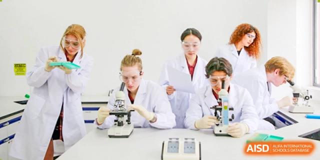Three students in lab coats and safety glasses looking at papers and using microscopes in a science class at Canadian International School of Hefei.