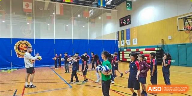 A group of students gathered around a student holding a soccer ball inside a brightly lit gymnasium with flags and a scoreboard at Canadian International School of Hefei.
