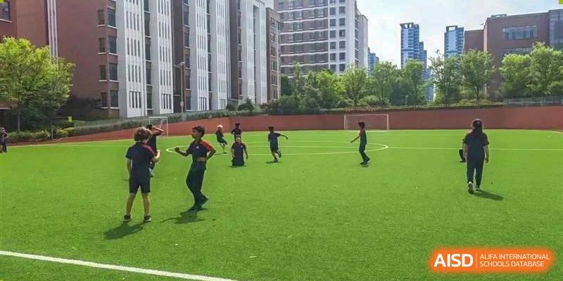 A group of students running and playing on a green outdoor sports field surrounded by tall buildings at Canadian International School of Hefei.