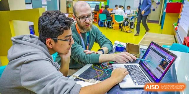 An adult mentor or teacher from Changchun American International School (CAIS) helping students with a project on a laptop.