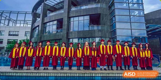 A line of graduating students from Changchun American International School (CAIS) in red and gold gowns posing in front of a modern school building.