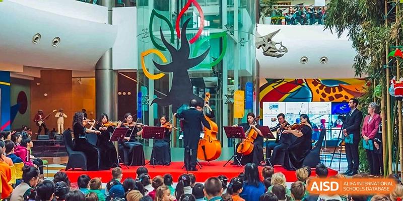 A small student orchestra from Changchun American International School (CAIS) performing on a stage with a modern sculpture backdrop.
