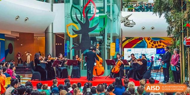 A small student orchestra from Changchun American International School (CAIS) performing on a stage with a modern sculpture backdrop.