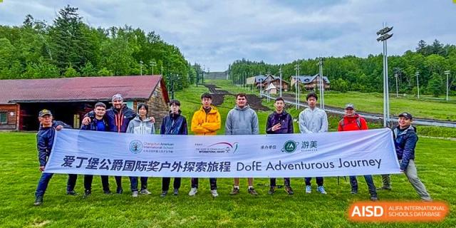 A group of students and staff from Changchun American International School (CAIS) holding a banner for the 'DoE Adventurous Journey' during an outdoor trip, with a small village in the background.