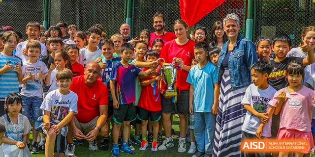 A group of students and teachers from Changchun American International School (CAIS) celebrating a sports victory, holding a large gold trophy.