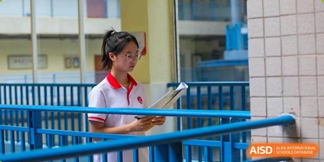 A female student in the distinctive red and white uniform stands in a school hallway, reading a book intently, highlighting academic focus at Guiyang New World School.