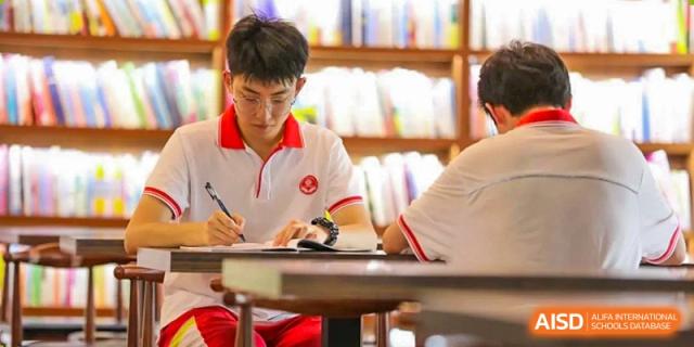 A male student in a white polo shirt with red trim is concentrating while writing notes at a desk in the school library or study area at Guiyang New World School.