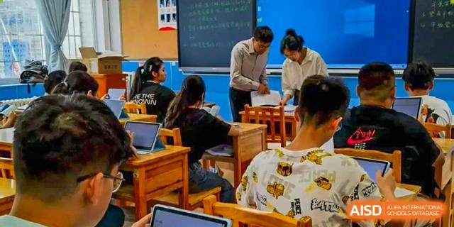 A teacher stands beside a student, reviewing papers and offering guidance to her in a classroom with a dark chalkboard background at Guiyang New World School.