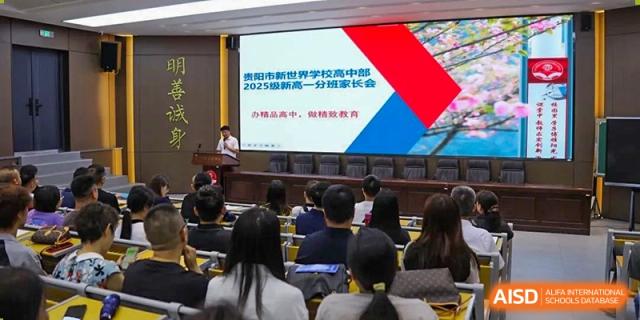 An auditorium where students and adults are seated, facing a large screen displaying Chinese text about a 2025 meeting, with a speaker at a podium. The school assembly is held at Guiyang New World School.