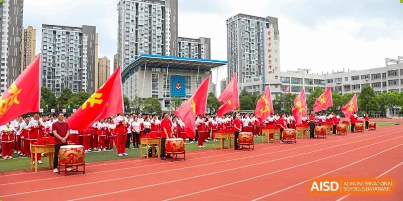 Students in white and red uniforms performing with large traditional drums on a red track in front of a modern school building with tall city buildings behind it. The event is at Guiyang New World School.