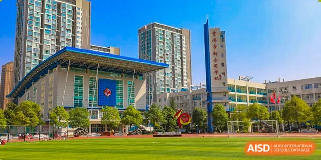 A wide exterior view of a modern school campus featuring a large blue and white administrative building, a green sports field, and tall urban towers. The central campus is Guiyang New World School.