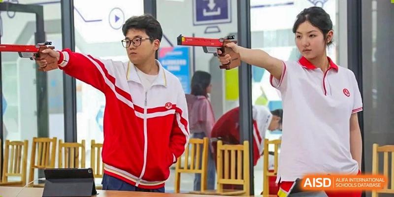 A student participating in a marksmanship activity, wearing a red and white jacket and aiming a practice gun in a classroom setting at Guiyang New World School.