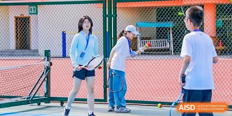 Two female students practice tennis on an outdoor court, holding rackets and preparing to hit the ball, demonstrating sports participation at Guiyang New World School.