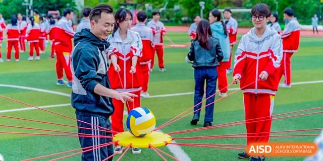 A group of students and a teacher participate in a fun physical education activity involving a volleyball on a spider web-like platform on the sports field at Guiyang New World School.