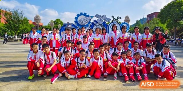 A large group of students in matching red and white uniforms are sitting together and smiling for a group portrait outdoors, celebrating school spirit at Guiyang New World School.