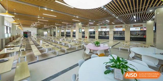 A bright, spacious school cafeteria with many long, empty tables and benches, modern wooden ceiling accents, and a round table in the foreground Guiyang New World School