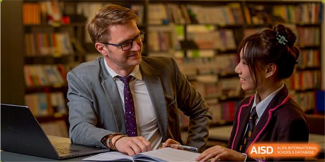 A male teacher wearing a suit and tie smiling while working at a desk surrounded by bookshelves at Haileybury Schools, Tianjin.