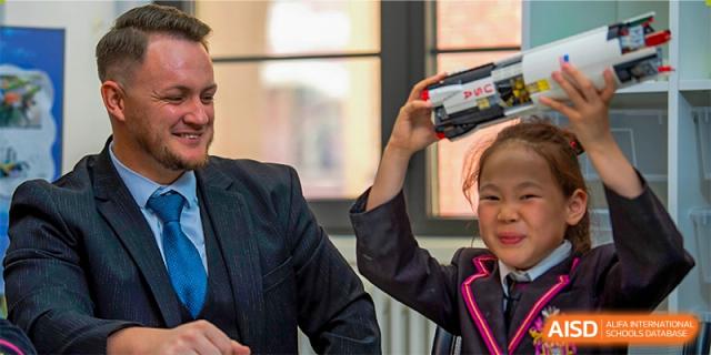 A teacher interacting with a male student who is holding a small electronic device or camera at Haileybury Schools, Tianjin.