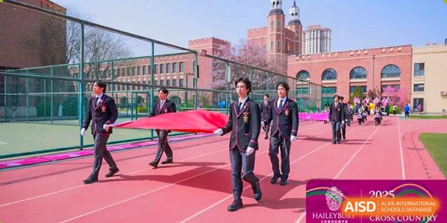 Teachers or staff in dark suits carrying a large red banner across the sports field at Haileybury Schools, Tianjin.