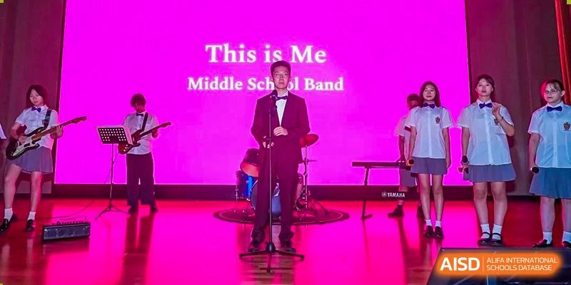 A student dressed in a black suit standing on stage in front of a neon sign that reads "This is Me Middle School Band" at Haileybury Schools, Tianjin.