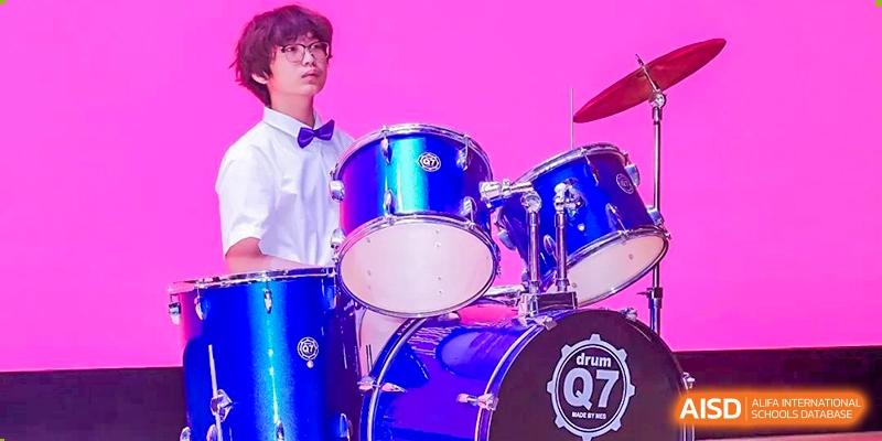A male student in a white shirt and bow tie sitting in front of a bright blue drum set during a performance at Haileybury Schools, Tianjin.
