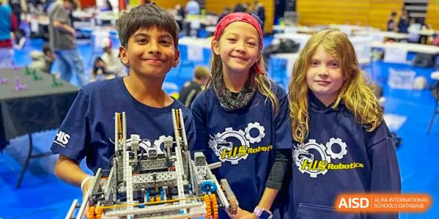 A group of smiling students from Hangzhou International School wearing team shirts and posing with a complex robotics build.